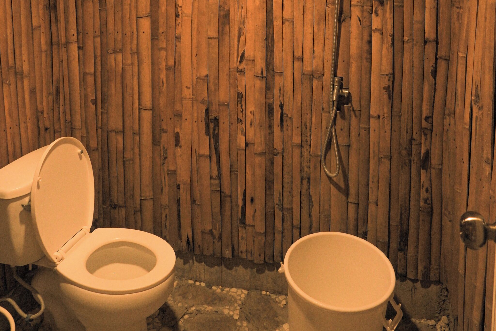 Une salle de bain avec un mur en bambou, des toilettes et un seau.