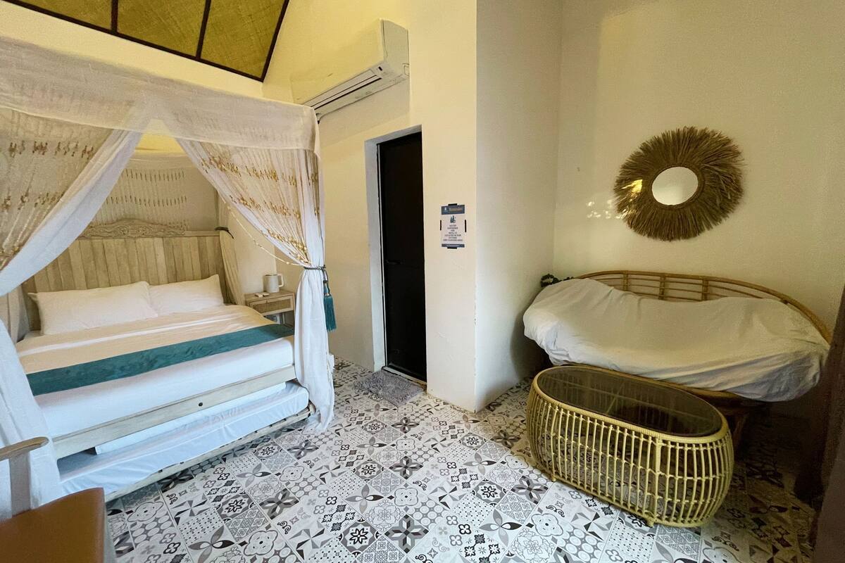 A bedroom with a bed, a bedside table, a bedside lamp, a wall-mounted mirror, a decorative wall piece, and a woven basket.