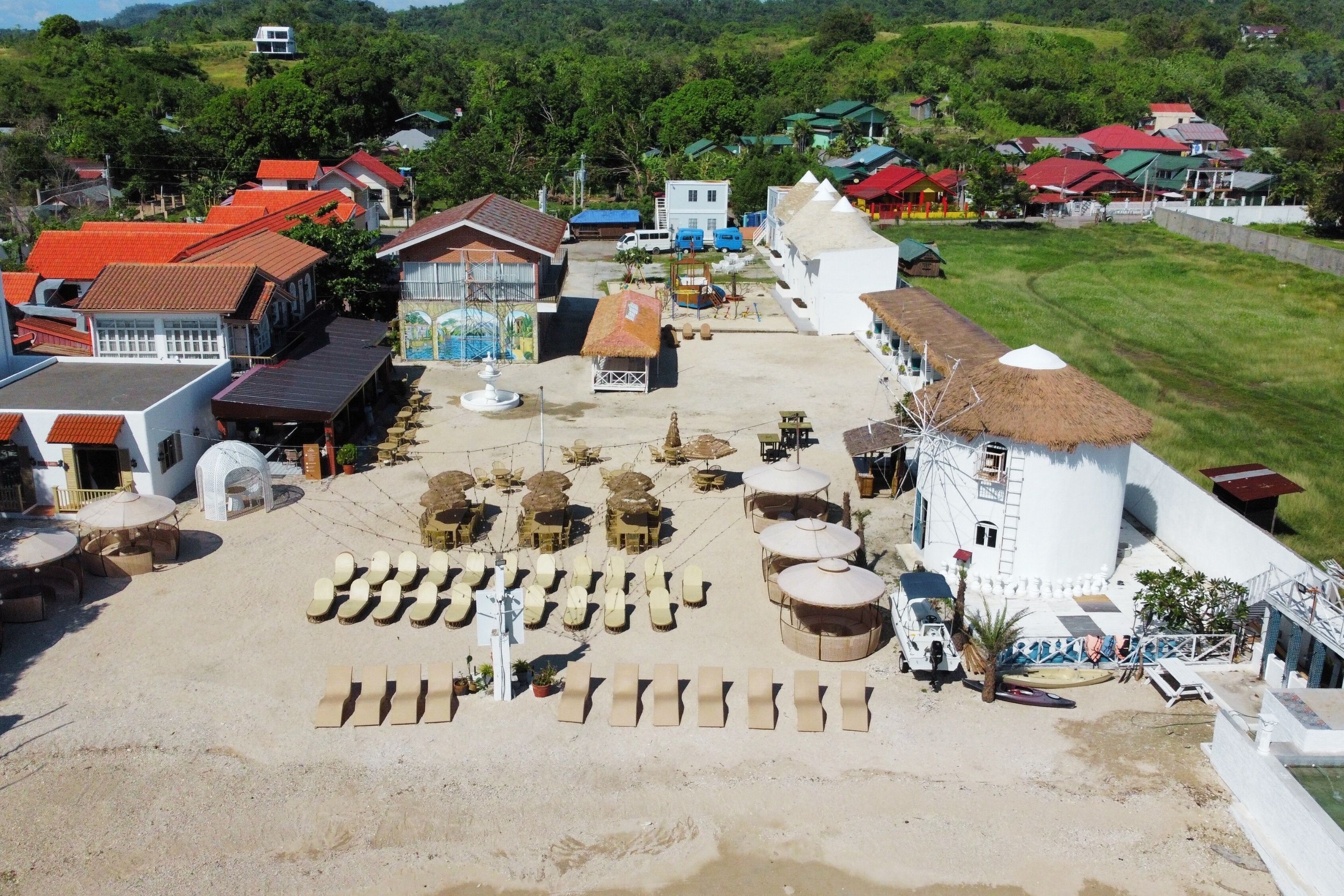 An aerial view of a coastal village with thatched-roof buildings, a sandy plaza with seating, and a white structure with a conical roof.