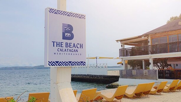 A beach sign for 'The Beach Calatagan, Mediterranean' with lounge chairs and a building in the background. - The Beach Calatagan Mediterraneo powered by Cocotel (Calatagan)
