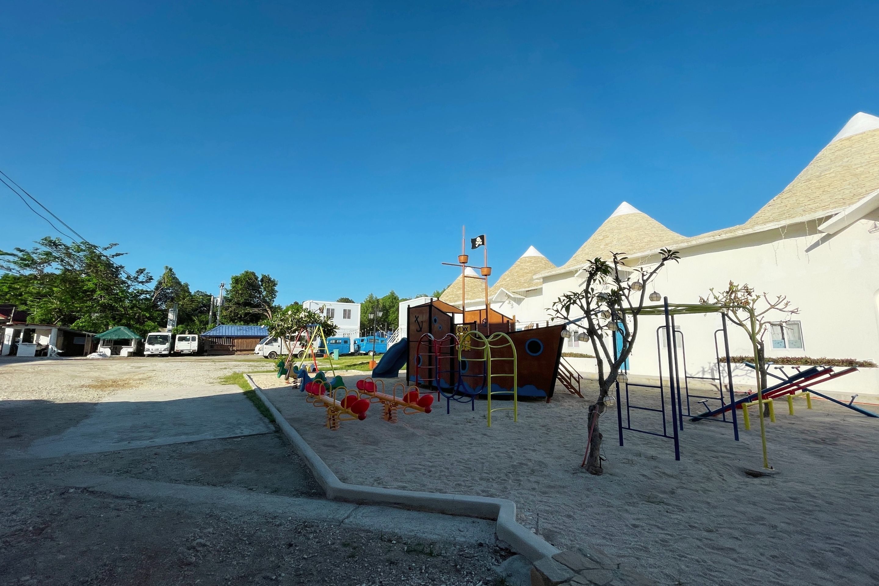 A playground with various play structures, including a seesaw and climbing frames, set against a backdrop of buildings with pyramid-shaped roofs.