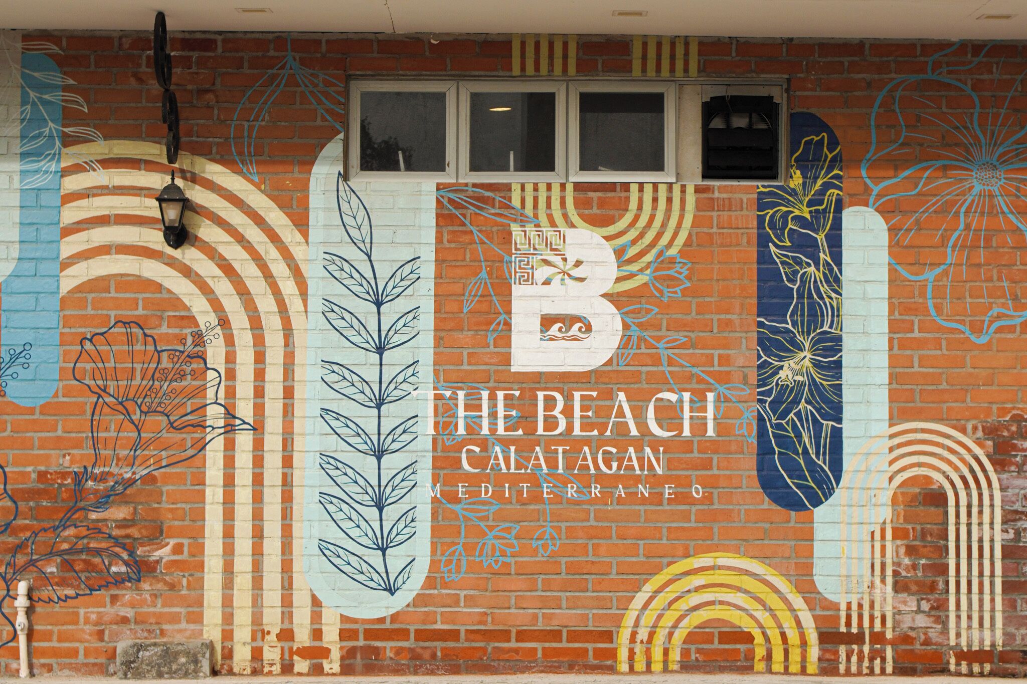 Una pared con un mural de una escena de playa, incluyendo un faro y olas, y el texto 'THE BEACH CALATAGAN MEDITERRANEO'.