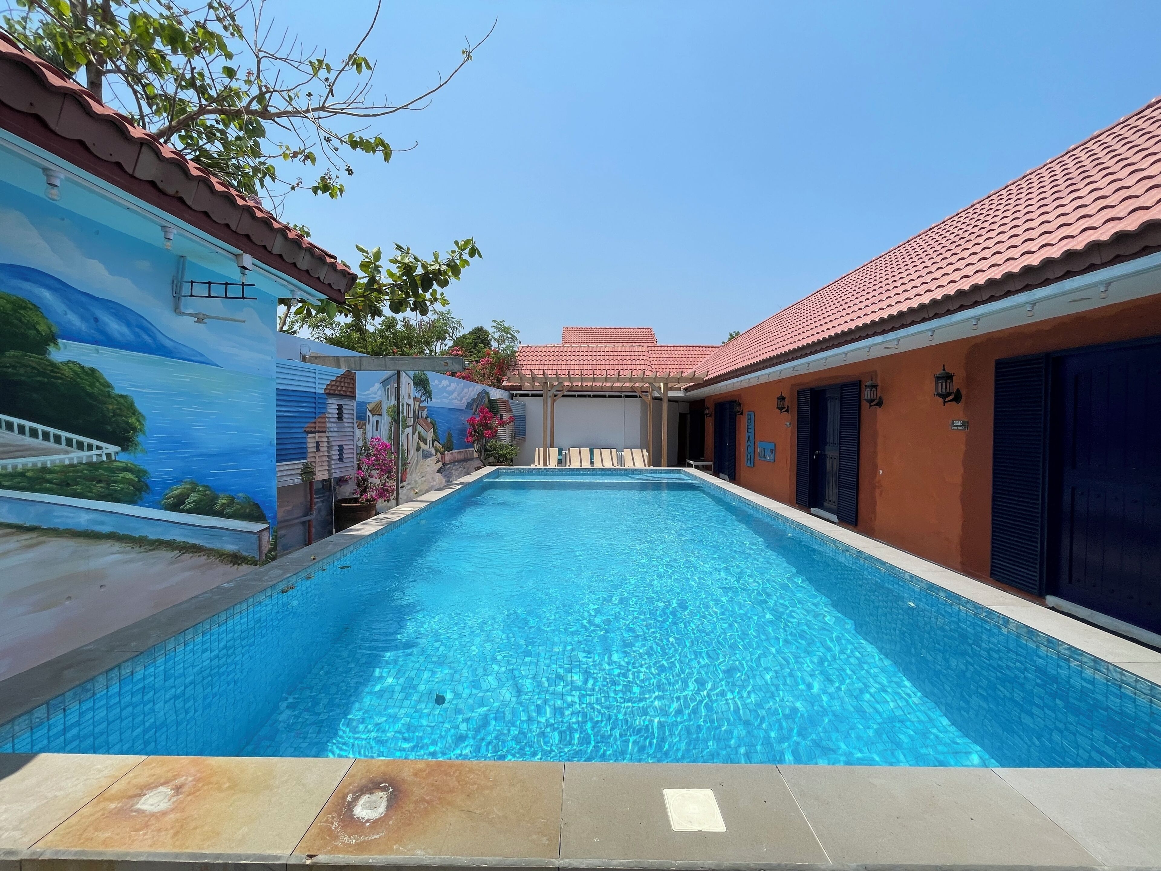 A swimming pool with a mural on the adjacent wall, a covered seating area, and a building with a red-tiled roof.
