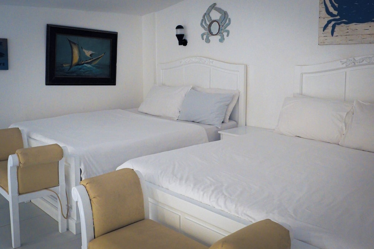 A room with two beds, each with white bedding and headboards, and two chairs with beige cushions.