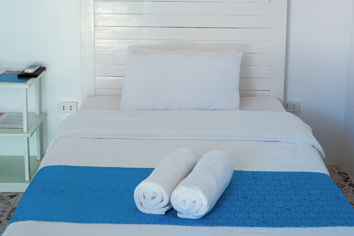 A neatly made bed with white bedding and two rolled towels, a white nightstand with a remote control, and a blue patterned bedspread.