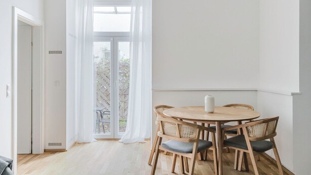 A dining area with a round wooden table and four chairs, a white vase on the table, and a view through a large window with sheer white curtains. - Blueground | Leopoldstadt, furniture & decor (Vienna)