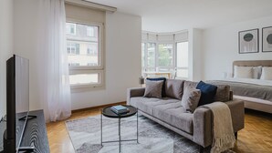 Un salon moderne avec un canapé, une table basse et un téléviseur. On y trouve des fenêtres avec des rideaux, un lit avec une tête de lit et des tableaux encadrés accrochés au mur.