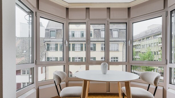Une pièce d’angle avec une table ronde et quatre chaises, de grandes fenêtres donnant sur des immeubles et un plancher en bois.