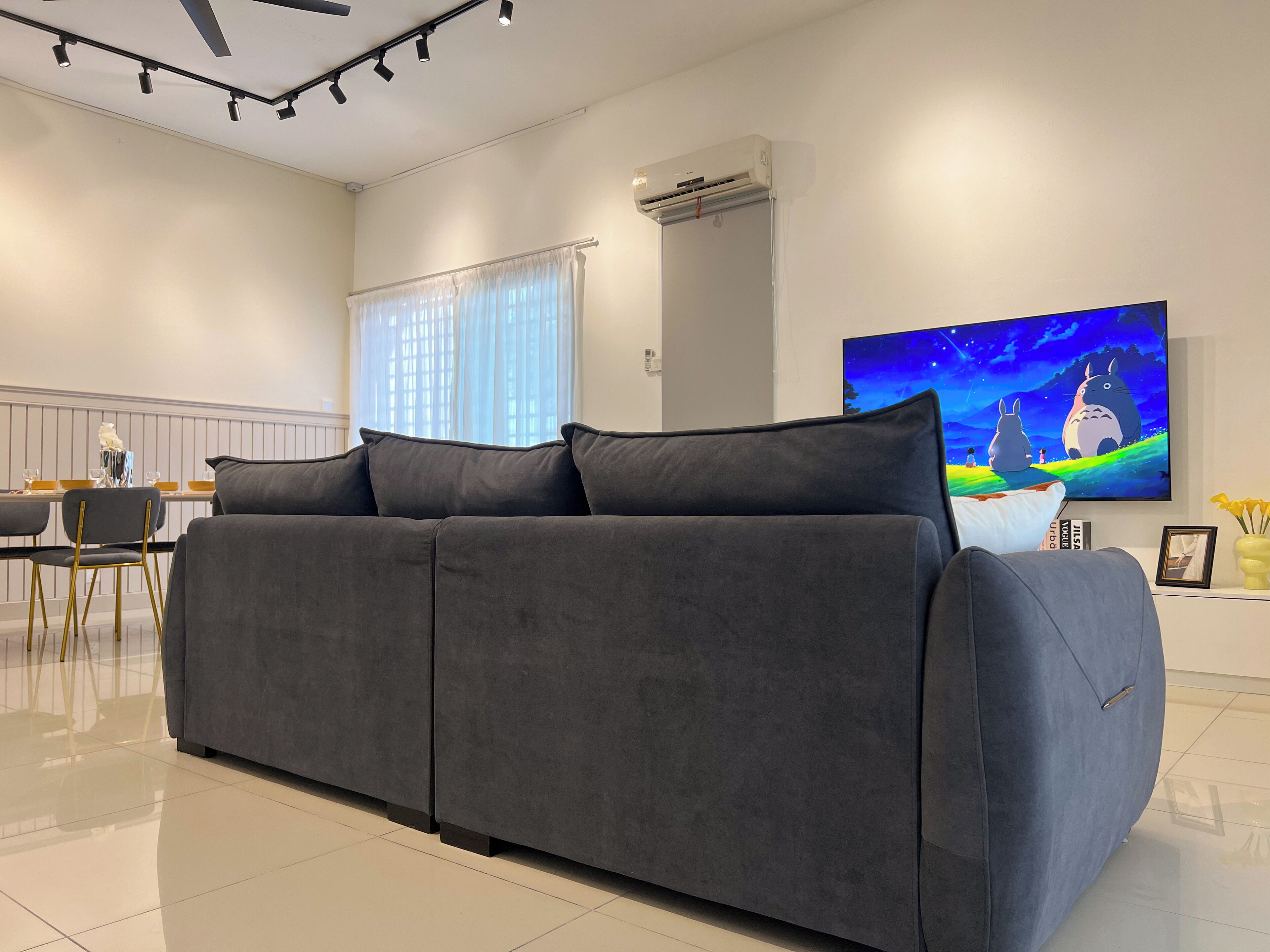 A modern living room with a grey sofa, a dining table with chairs, a large flat-screen TV, and a window with sheer curtains.