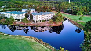 Um hotel com vários andares, com uma área de água refletora na frente, rodeado por vegetação e um campo de golfe.