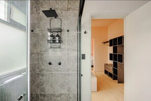 A modern bathroom with a glass shower enclosure, a built-in shelf with toiletries, and a view into a bedroom with a wooden floor and built-in shelving.