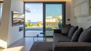 En moderne stue med en grå sofa, et lille bord og en stor skydedør, der fører ud til en balkon med udsigt over havet og bygninger.