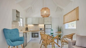 A modern kitchen with white cabinets, a dining area with wicker chairs, a blue armchair, and a round coffee table. - Villa Artemis Periyali (Zakynthos)
