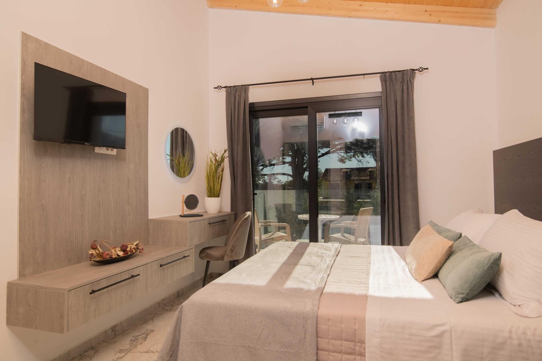 A modern bedroom with a bed, a TV mounted on the wall, a desk with a chair, and a balcony with a view of the outside.
