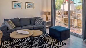 Una sala de estar moderna con un sofá gris, dos mesas auxiliares, una lámpara y vistas a un patio de piedra a través de grandes ventanales.