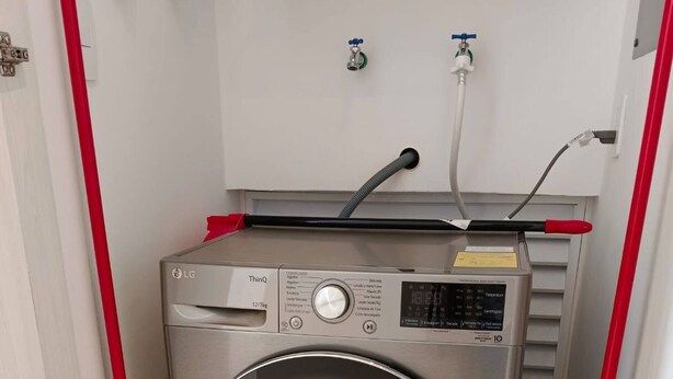 A compact, stainless steel front-loading washing machine in a small laundry room with a red mop and broom, a water heater, and a water tap. - Beautiful 1 Bedroom Aldea Zama en Tulum (Tulum)