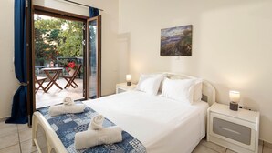 Ein ordentlich bezogenes Bett mit einer gemusterten Decke und zwei Handtüchern, ein Bild an der Wand und ein Blick ins Freie durch eine Glastür.