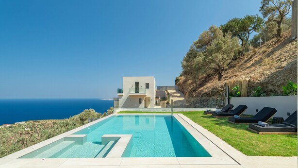 Un espace piscine moderne offrant une vue dégagée sur la mer, une colline parsemée d’arbres et un bâtiment en arrière-plan.
