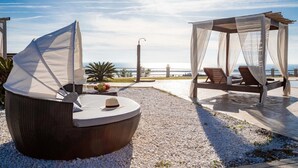 Een strandlounge met een rieten stoel en een hemelbed, beide met witte kussens en een hoed op de stoel.