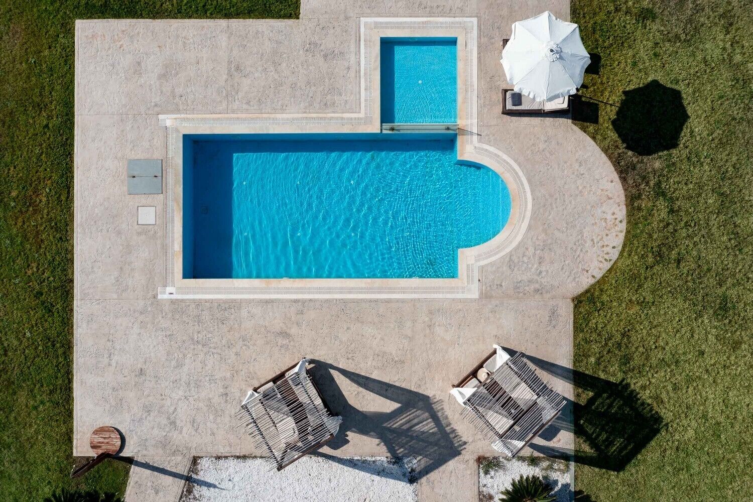 Vista aérea de una piscina de forma rectangular y borde curvo, rodeada por un suelo de hormigón. Hay dos sillas reclinables y una sombrilla blanca en el suelo.