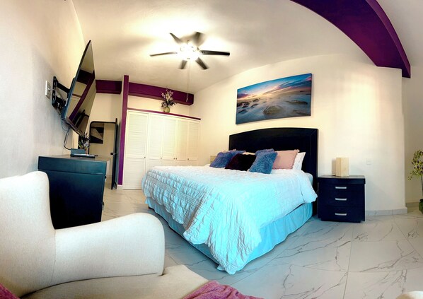 A bedroom with a bed, a desk, a chair, a ceiling fan, a painting, and a wardrobe.
