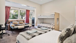 Ein Schlafzimmer mit einem großen Bett, einem kleineren Bett, einem Schreibtisch und einem Sessel. Ein Fenster mit Vorhängen bietet Blick auf Bäume.