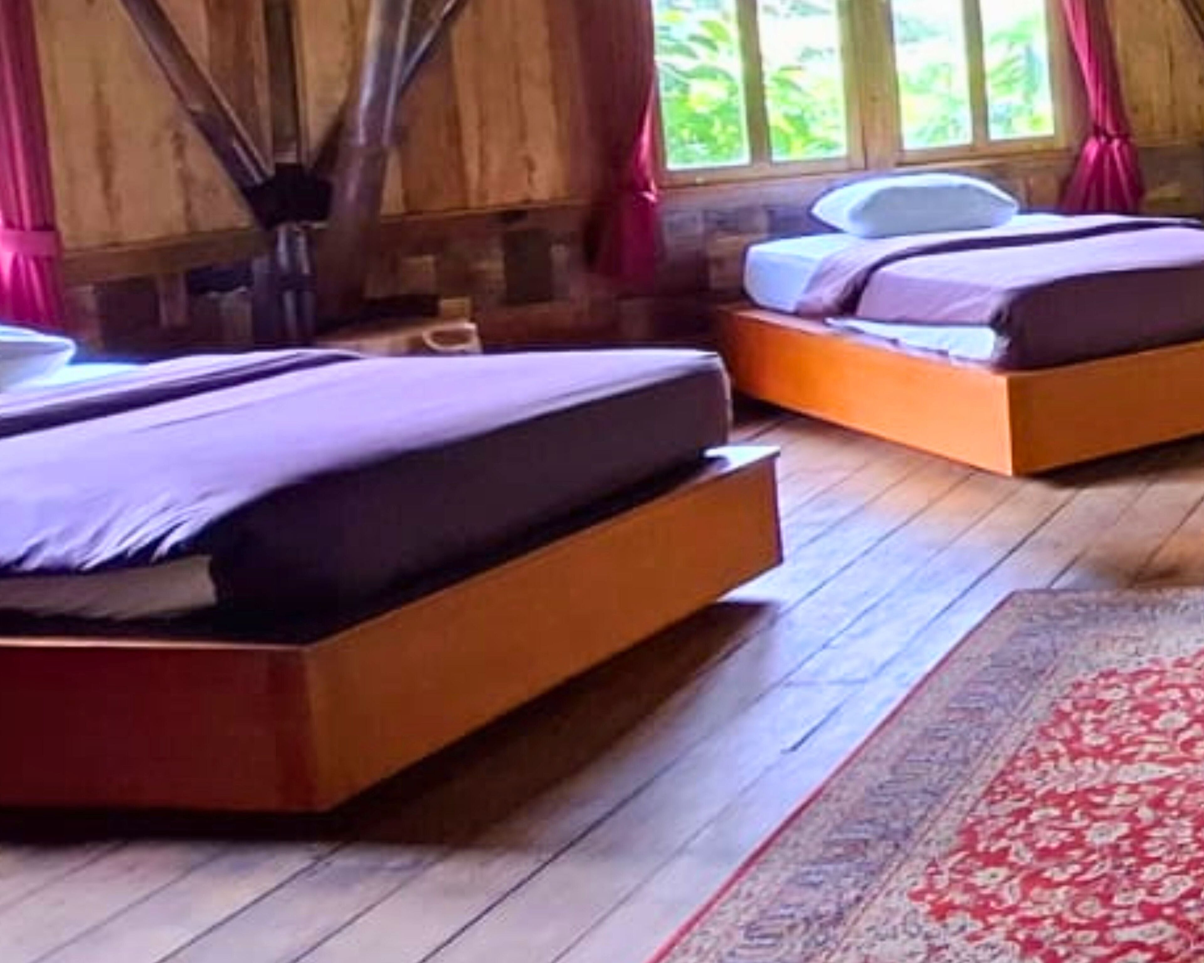 A room with two beds, wooden floors, and a large window with curtains.