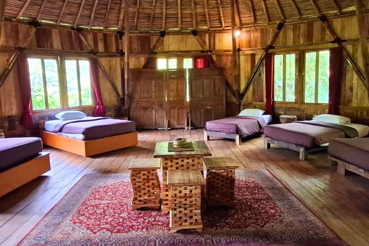 A rustic wooden cabin with four beds arranged around a central wooden table, large windows, and a thatched roof.