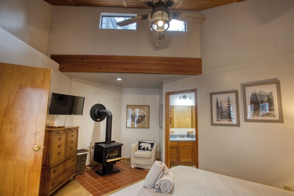 Un dormitorio con techo de madera, una cama, un aparador de madera, un televisor, una chimenea, una silla y cuadros enmarcados en la pared.