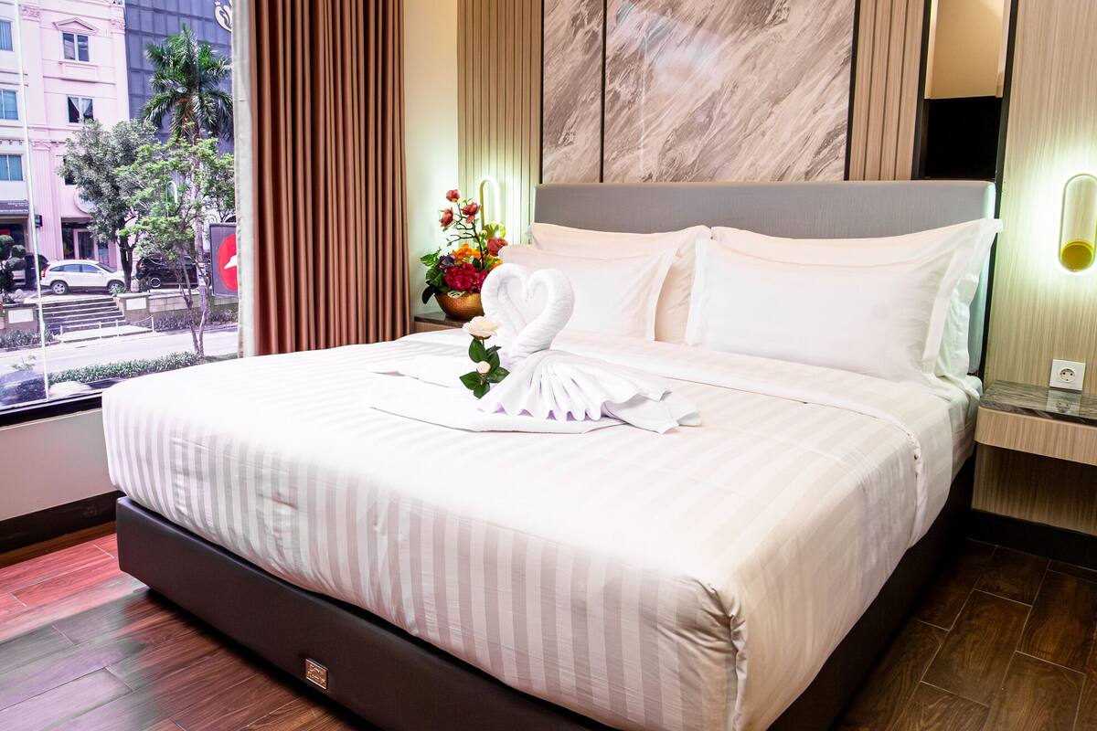 A hotel room with a large bed, a window with curtains, a vase of flowers, and a view of the street outside.