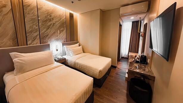 A hotel room with two beds, a TV, a desk, and a trash bin. - d'primahotel PIK Jakarta (Jakarta)