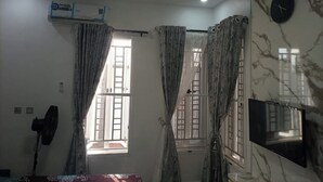 A room with a ceiling fan, air conditioning unit, two windows with curtains, a wall-mounted TV, and a clock.