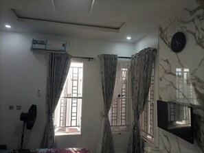Une pièce dotée d’un ventilateur de plafond, d’un appareil de climatisation, de deux fenêtres avec rideaux, d’un téléviseur fixé au mur et d’une horloge.