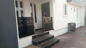 A building with a white exterior, black-framed windows, and a metal gate. There are steps leading to the entrance and a trash bin to the side.