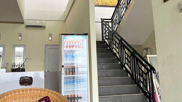 A hotel lobby with a reception desk, a refrigerator stocked with beverages, a wicker chair with a floral cushion, and a staircase with a black railing. - Djuragan Kamar Macan Kumbang (Palembang)