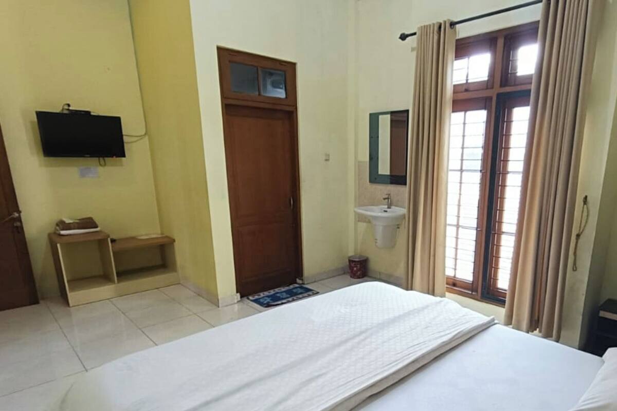 A hotel room with a bed, a small bedside table, a TV mounted on the wall, a window with curtains, and a sink with a mirror.