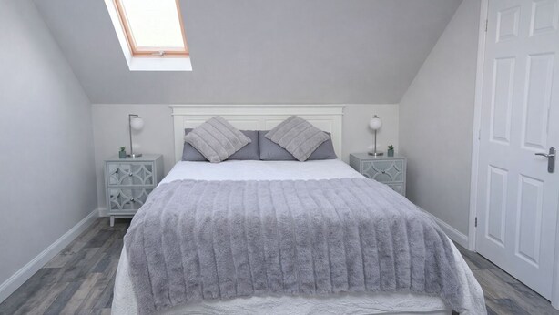 Een slaapkamer met een schuine plafond, een dakraam, een bed met een zachte grijze deken, twee nachtkastjes met lampen en een witte deur.