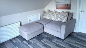 Een moderne woonkamer met een grijze modulaire bank, een bijpassend poefje, een witte radiator, en een schilderij met een strandthema aan de muur.