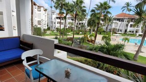Un balcón con un sofá azul, una silla blanca y una mesa de vidrio. Se ve una piscina, palmeras y un edificio con varios balcones.
