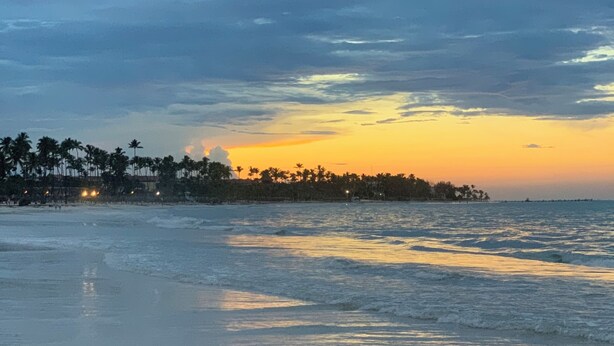Una playa con aguas tranquilas que reflejan el cielo, una costa lejana con palmeras y un horizonte despejado.