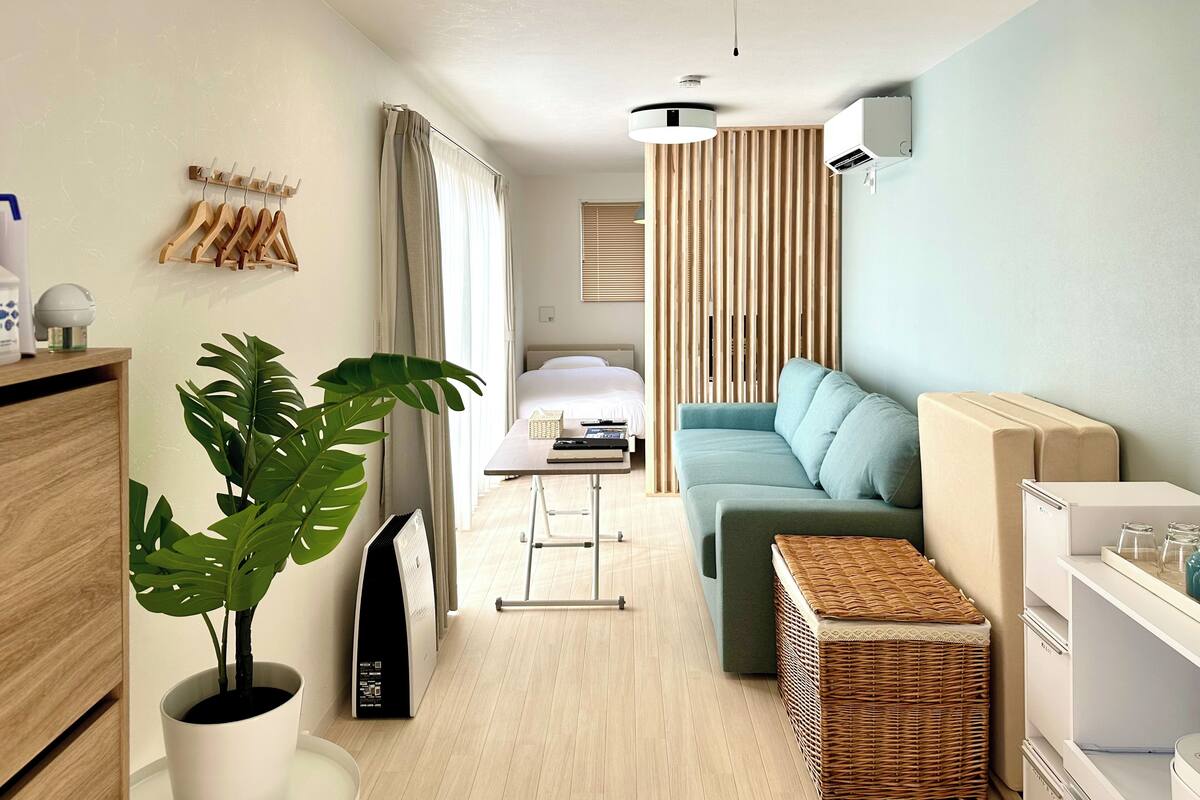 A modern hotel room with a bed, a sofa, a dining table, and a potted plant.