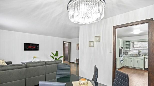 Ein modernes Wohnzimmer mit grauem Sofa, einem Esstisch mit Stühlen, einer Kristallleuchtre und einer durchgehenden Tür einsehbarer Küche.