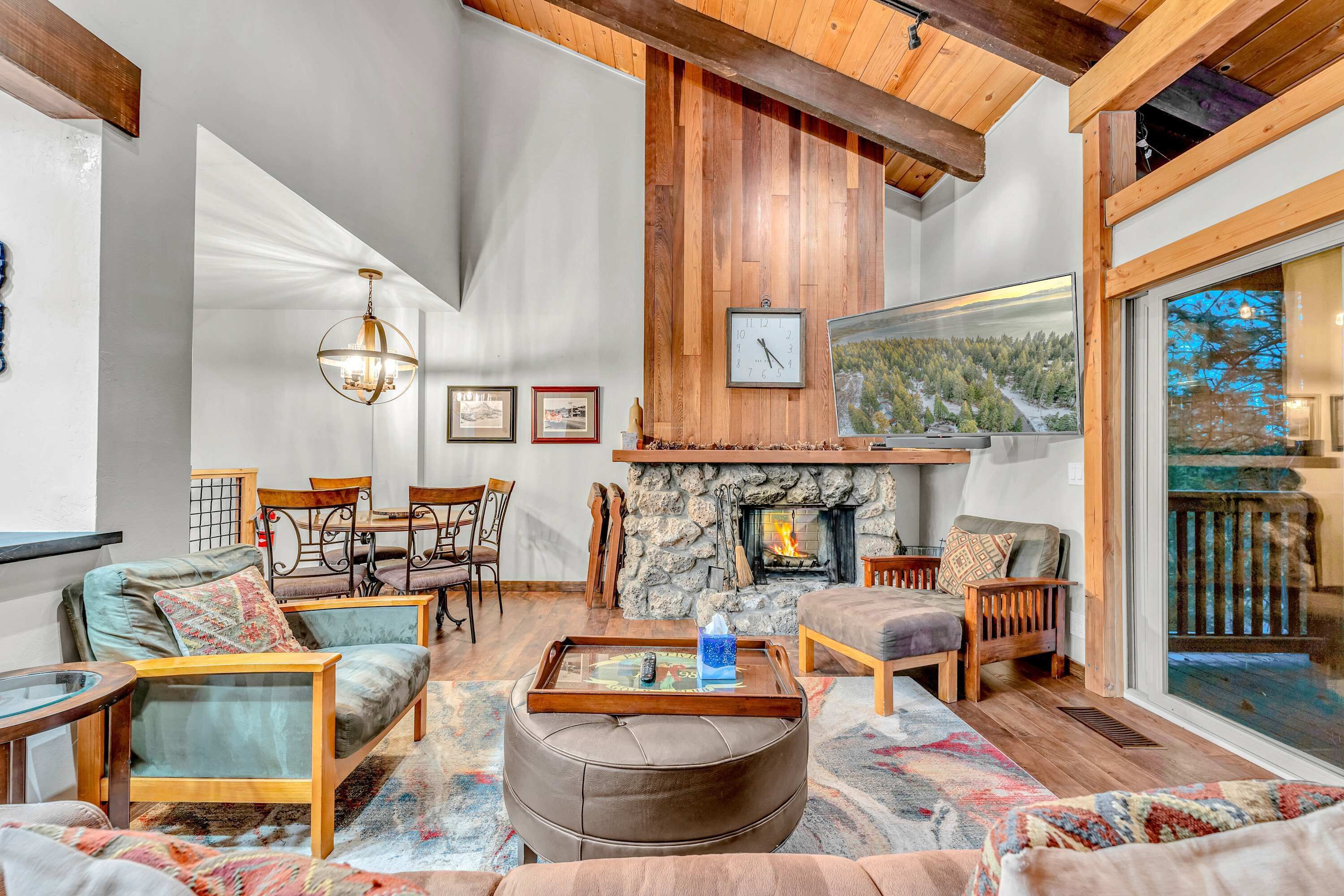 A cozy living room with a stone fireplace, wooden ceiling beams, and a large window offering a view of the outdoors.