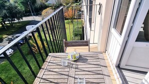 A wooden balcony with a table set for two, overlooking a green garden and a street.