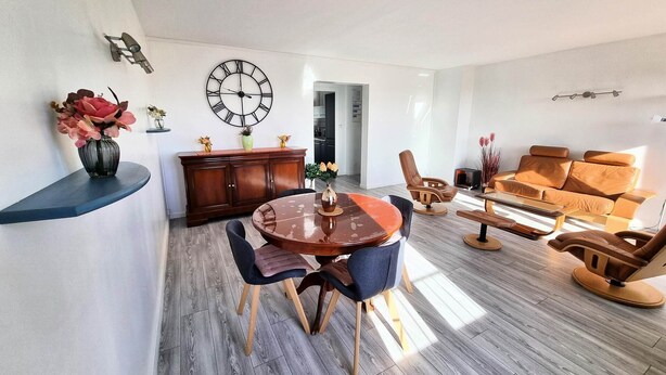 A modern living room with a round wooden dining table, two chairs, a sofa, and a coffee table. There is a large wall clock and a small shelf with flowers.