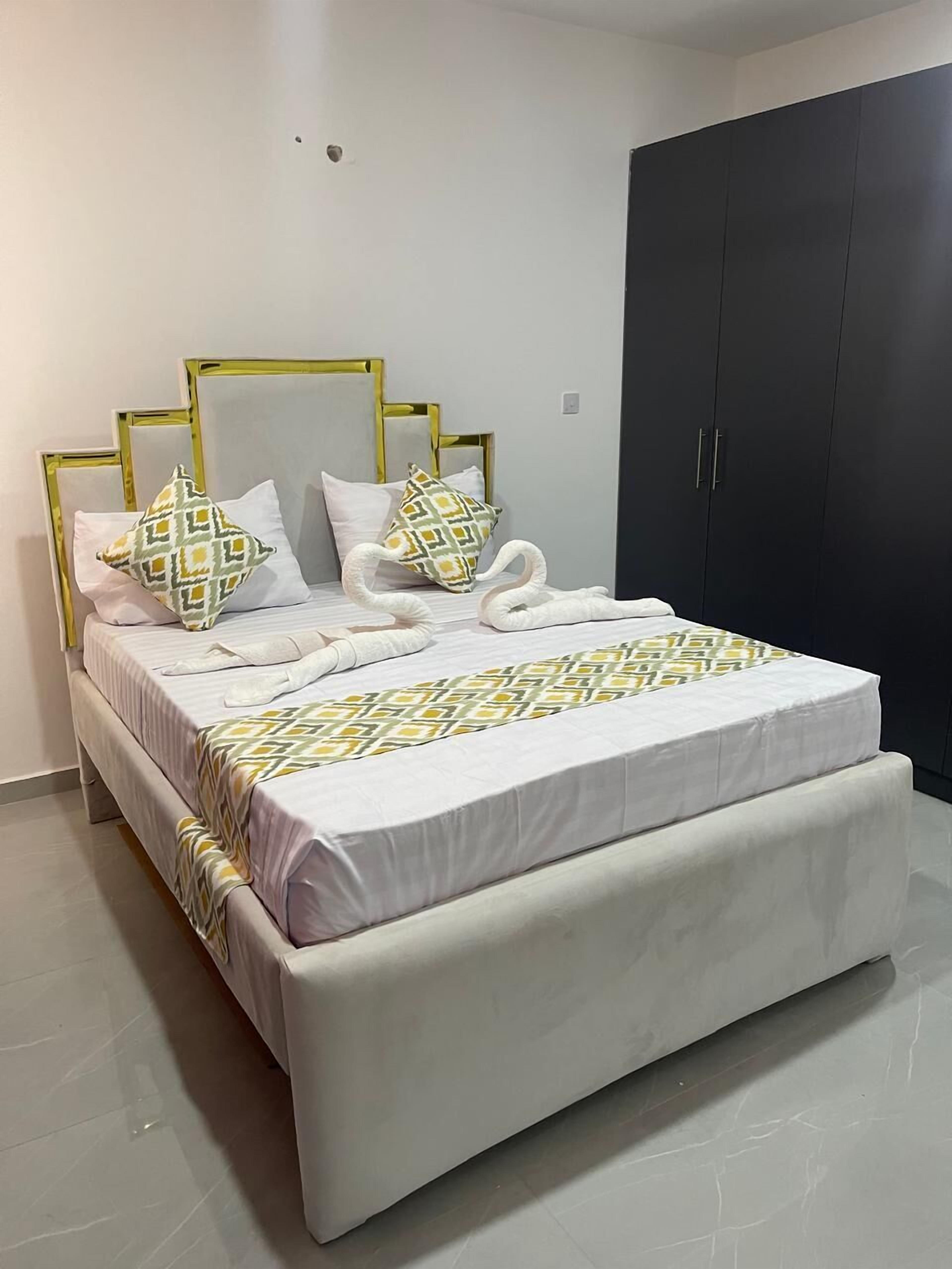 A neatly made bed with a patterned bedspread and pillows, a dark wardrobe, and a plain wall.