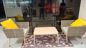 Uma área de estar com cadeiras de vime e uma pequena mesa, um tapete estampado e um painel de vidro refletindo o interior.