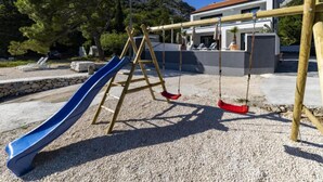 Un parque infantil con tobogán y columpios, con un edificio moderno y árboles de fondo.