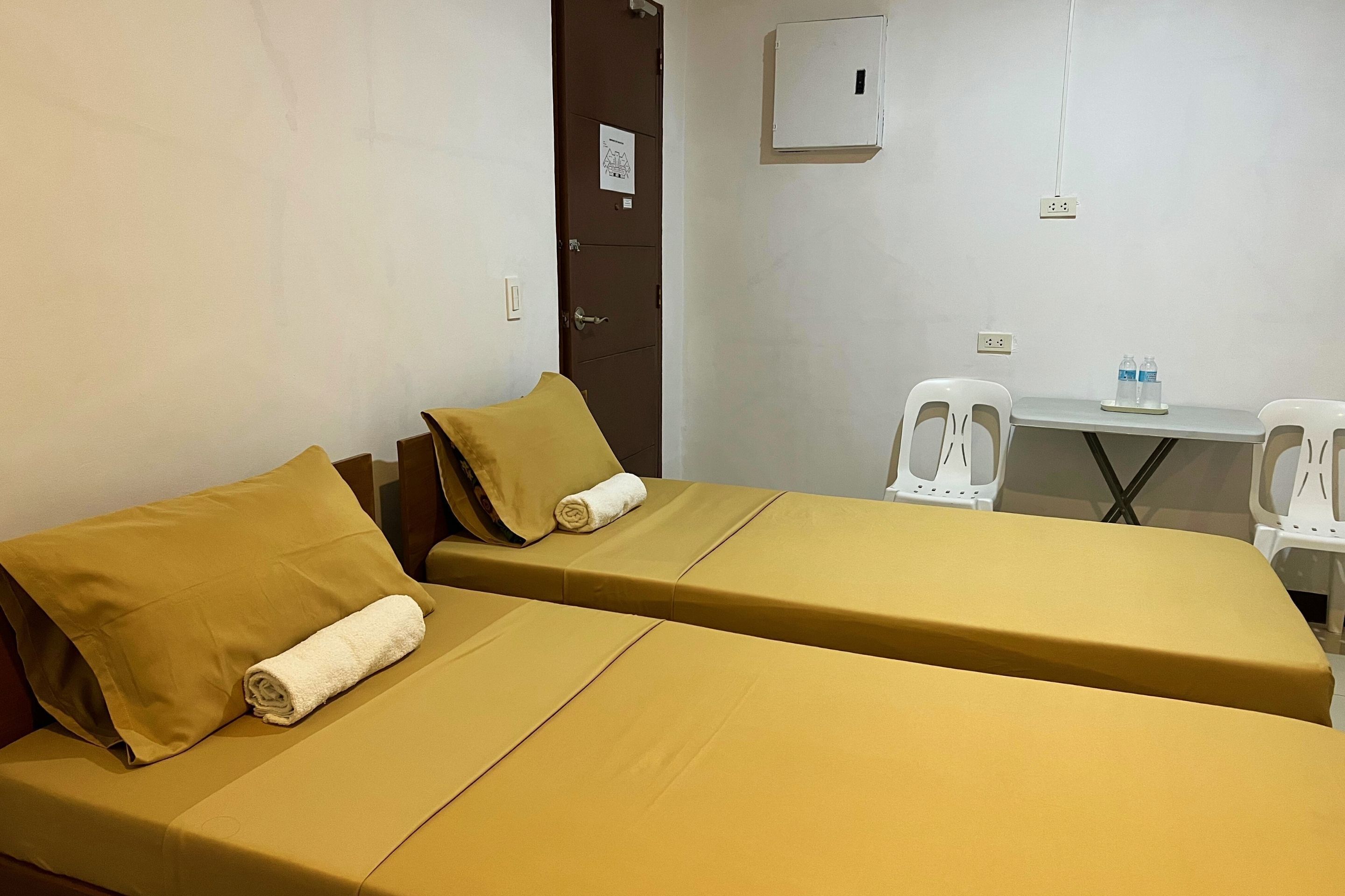 A hotel room with two beds, each with a yellow bedspread and pillows, a small table with a water bottle, and a whiteboard on the wall.