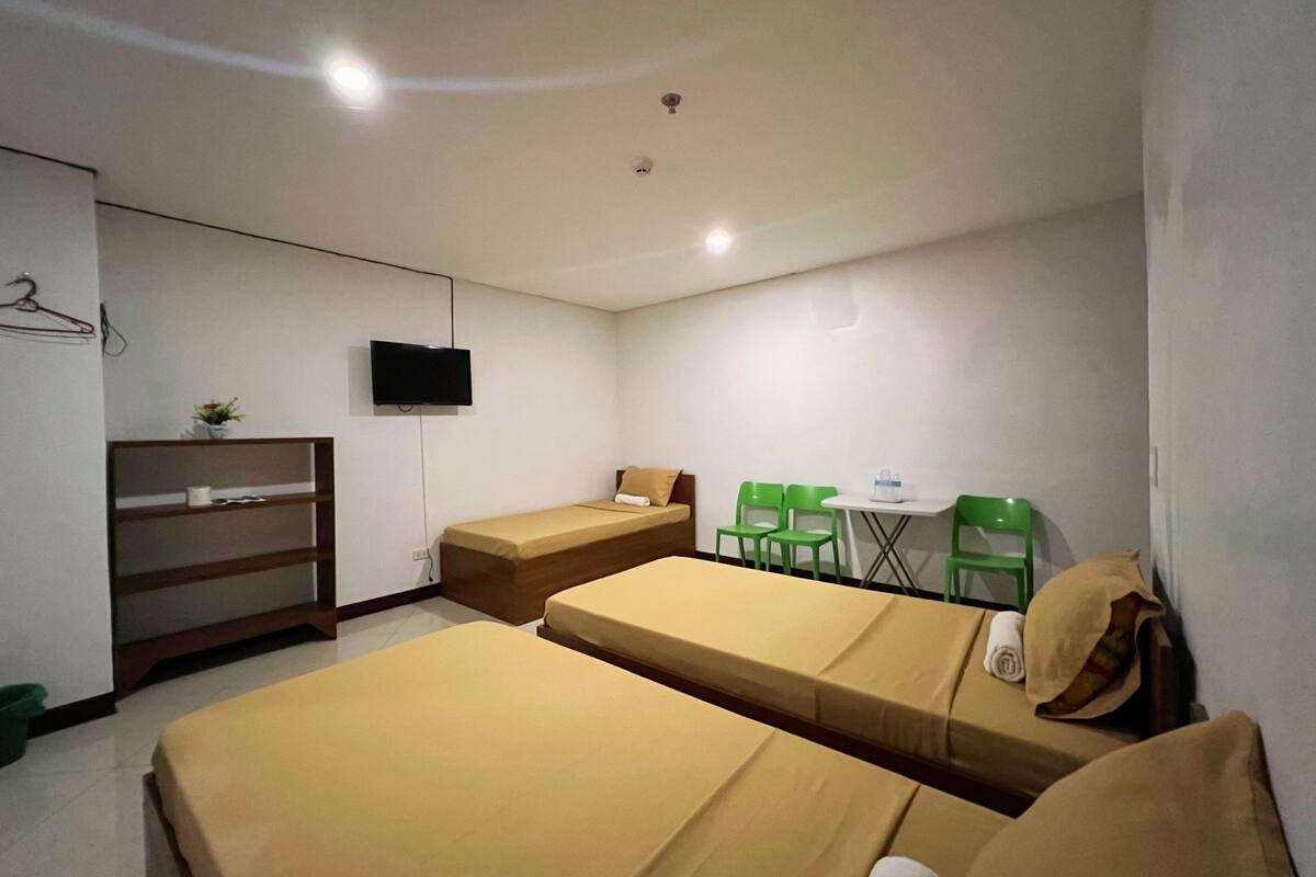 A hotel room with two single beds, a TV on the wall, a small table, and two green chairs.